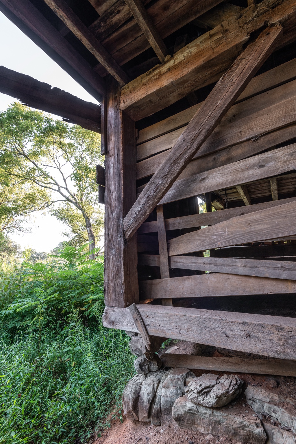 Timber post, beam and diagonal brace above a stone foundation, with weathered wooden siding and trees alongside the building
