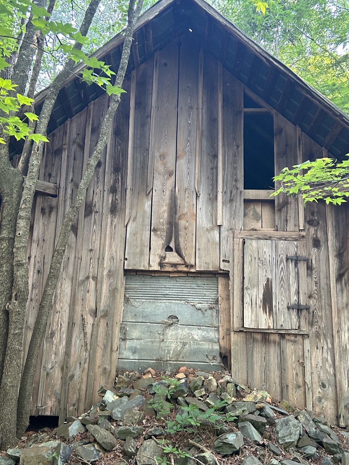 Weathered wooden outbuilding among trees, with vertical plank siding, a shuttered opening and stone rubble at its base
