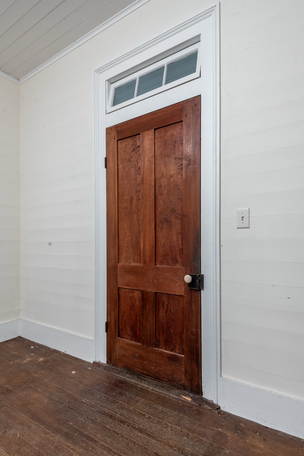 Wooden four-panel door with a white knob and three-pane transom window, surrounded by white plank walls and wooden floorboards